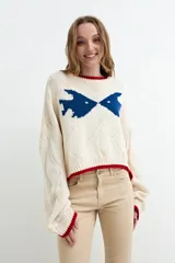 Sweater tejido color marfil con diseño de peces en azul en el pecho y detalles de ribetes rojos en el cuello, puños y ruedo. Presenta mangas largas abullonadas y textura de rombos en relieve.