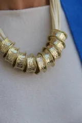 Collar de diseño artesanal compuesto por múltiples tiras de cuero en color crudo y piezas cilíndricas de metal dorado con textura martillada.