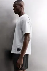 Camiseta de ajuste relajado (relaxed fit) color terracota, confeccionada en tejido técnico. Presenta cuello redondo, mangas ranglán cortas y un parche negro con logo en el bajo.