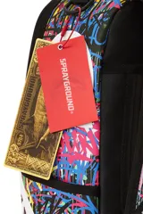 Mochila Sprayground con estampado de graffiti multicolor y diseño de boca de tiburón con dientes blancos. Cuenta con bolsillo frontal con cremallera, bolsillos laterales, bolsillo oculto con cremallera, compartimento para gafas de sol, acolchado ergonómico en la parte posterior, correas ajustables, cierres dorados, placa metálica con logo, compartimento para laptop y espalda ergonómica con malla.