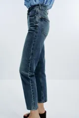 Jeans de mujer, tiro alto, corte Mom Fit con pierna slim y largo tobillero. Denim azul medio con efecto desgastado y lavado. Presenta cinco bolsillos y cierre frontal con cremallera y botón.