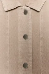 Sobrecamisa de mujer color beige claro, con cuello solapa, manga larga y bolsillos de plastrón en el delantero. Presenta bajo asimétrico con aberturas laterales y cierre frontal de botones.