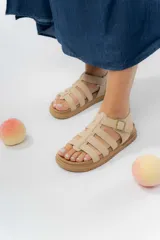 Sandalia estilo gladiadora de color beige, confeccionada en material gamuzado sintético. Presenta múltiples tiras anchas que sujetan el pie, ajuste lateral con hebilla metálica y suela plana antideslizante de color marrón.