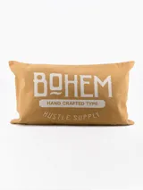 Funda de almohadón rectangular color beige con estampa tipográfica en blanco con la palabra "BOHEM" y la frase "HAND CRAFTED TYPE HUSTLE SUPPLY".