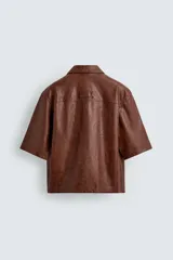 Sobrecamisa de corte relajado confeccionada en tejido efecto piel, con cuello solapa, manga corta, bolsillo plastrón en el pecho y cierre frontal con cremallera.