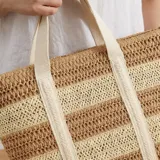 Bolso tipo tote confeccionado en paja tejida con diseño de rayas horizontales en tonos beige y marrón. Cuenta con doble asa de hombro de lona clara y cierre superior.