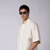 Camisa de manga corta color marfil, confeccionada en 100% algodón con calce boxy, cuello camisero, cierre frontal de botones y diseño con bordado sutil en la parte inferior.