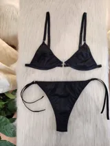 Conjunto de bikini color negro con textura acanalada. El corpiño es de estilo triangular con aros y detalle de argolla metálica en el centro. La bombacha es de corte colaless con tiras laterales ajustables y detalles de argollas metálicas.