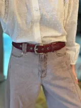 Cinturón de cuero bordó con detalle de costura entrelazada en los bordes, hebilla metálica plateada y ojales reforzados.