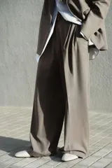 Pantalón sastrero de corte recto y perneras amplias, color marrón, con pinzas frontales, trabillas ajustables con botones en la cintura y raya marcada en el centro de las piernas.