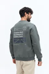 Buzo de algodón y poliéster color verde musgo, con cuello redondo y puños acanalados. Presenta un parche estampado rectangular con texto y un logo circular en el pecho.