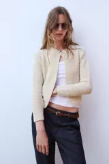Chaqueta de punto de hilo metalizado color beige con cuello redondo, manga larga y cierre frontal con un botón dorado.
