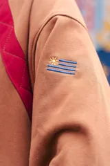 Buzo amarillo de corte holgado con mangas raglán en color azul, detalle de costuras acolchadas en los hombros y bordado de sol en el centro del pecho. Presenta puños y cuello acanalados con rayas azules.