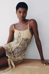 Vestido corto semitransparente tipo corsetero color beige, confeccionado con encaje y detalles de varillas interiores que estilizan la figura. Presenta escote redondo, tirantes finos ajustables y bajo asimétrico con una lazada lateral de satén.