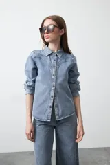 Camisa de jean color azul claro con tachas metálicas en el canesú y a lo largo de las costuras frontales. Tiene mangas largas con puños abullonados y cierre frontal con botones.