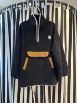 Buzo negro tipo corderito con cuello alto y cierre parcial con cremallera. Tiene un bolsillo tipo canguro con apliques de pana color mostaza y un parche con letras en el pecho. El ruedo y los puños tienen ribete de color.