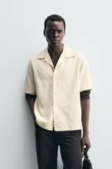 Camisa de corte relajado confeccionada en tejido de algodón, con cuello bowling y manga corta. Presenta un diseño con bordados geométricos a tono y cierre frontal de botones.