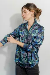 Camisa de seda con cuello clásico y cierre frontal de botones, de manga larga con puños abotonados. Presenta un estampado floral en tonos azules, verdes y rosados sobre fondo negro.