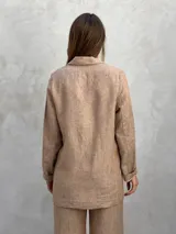Blazer de lino color beige con finas rayas verticales oscuras. Presenta solapas de muesca, dos botones frontales y bolsillos de parche en la parte inferior. El borde de las solapas y los puños tiene un ribete oscuro contrastante.
