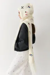 Bufanda con capucha integrada, confeccionada en tejido de punto grueso color crudo. Presenta bordados coloridos de figuras deportivas y cuenta con flecos en los extremos.