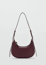 Bolso de hombro tipo hobo en color bordó, con diseño de media luna, cierre de cremallera y asa ajustable con hebillas metálicas.