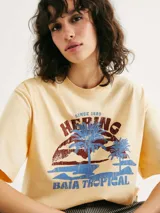 Camiseta color crema de corte amplio con estampado de paisaje costero enmarcado por columnas y la frase "Blue Horizon" en celeste.