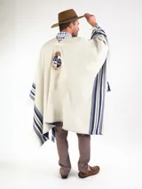 Poncho grueso color beige con detalles de rayas azules en los laterales y flecos en los bordes. Presenta el escudo bordado en la espalda y diseño liso en la parte frontal con escote en V.