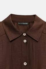 Camisa de corte recto confeccionada en tejido con mezcla de lino, color marrón. Presenta cuello solapa, cierre frontal con botones, mangas largas con puños y hombreras marcadas que aportan estructura.