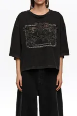 Remera corta de corte oversize color negro, confeccionada en mezcla de algodón. Presenta cuello redondo, mangas cortas amplias y un gran bordado frontal con diseño estilo etiqueta y texto.