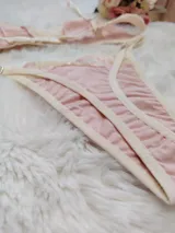 Conjunto de lencería compuesto por un bralette triangular y una tanga, confeccionados en algodón color rosado con detalles de encaje en tono crema. El bralette cuenta con breteles ajustables y el conjunto presenta terminaciones delicadas.