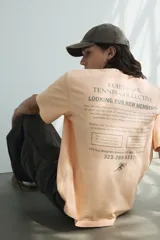 Remera color durazno de corte holgado con cuello redondo y estampado pequeño en el pecho con un tenista y texto.