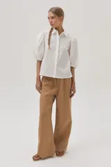 Camisa blanca de mangas abullonadas con puños elásticos, cuello clásico y cierre frontal con botones. Presenta detalles de bordado calado en el pecho.