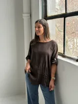 Blusa de lino color marrón chocolate, de corte holgado, con cuello redondo, mangas tres cuartos con trabilla y botón para sujetar remangadas, y detalle de alforzas verticales en el frente.