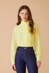Blusa de gasa color amarillo pastel, con cuello alto y lazo frontal tipo corbatín. Presenta mangas largas con puños abotonados y un diseño elegante y fluido.