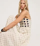 Vestido midi de algodón, color crema, con breteles finos. El corpiño presenta un diseño de crochet con motivos florales en azul marino, y la falda evasé tiene un estampado de pequeños íconos abstractos en azul.