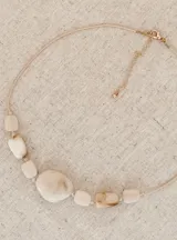 Collar de diseño artesanal compuesto por piedras irregulares en tonos crudos veteados, con una pieza central de mayor tamaño. Presenta un cordón beige ajustable y detalles en tono dorado.