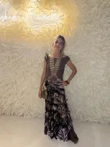Vestido largo de tul con estampado paisley en tonos marrones, negros y lilas. Presenta un diseño de cuerpo ajustado con cuello redondo y falda con volados en el ruedo. Incluye un detalle de arandela metálica en la cintura y una pieza de joyería dorada superpuesta en el pecho.
