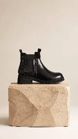 Bota tipo Chelsea de cuero negro, con paneles laterales elásticos y detalle de textura grabada en la parte trasera. Cuenta con suela track dentada y tirador en la parte superior.