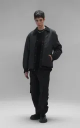 Campera tipo sobrecamisa de diseño oversize, confeccionada en microfibra opaca con patrón matelaseado geométrico. Presenta cuello, puños, cartera central y ruedo en gabardina de algodón, con cierre frontal mediante broches a presión. El diseño del ruedo simula una prenda superpuesta.
