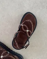 Sandalia de plataforma negra con tiras decoradas con tachas metálicas plateadas. Posee un diseño tipo ojota con ajuste de pulsera al tobillo mediante hebilla.