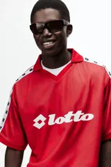 Camiseta de futbol de manga corta de la marca Lotto, en color rojo con un sutil estampado geométrico. Cuenta con cuello tipo polo abierto con escote en V y un calce holgado. Las mangas presentan una franja blanca lateral decorada con el logo de la marca repetido. El logotipo de Lotto destaca en blanco en el centro del pecho.