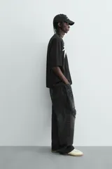 Camiseta negra de corte relaxed fit, cuello redondo y manga corta. Presenta un estampado frontal en blanco con efecto spray que simula dos figuras volando, y texto en un óvalo que dice "SUCCESS CHASER".