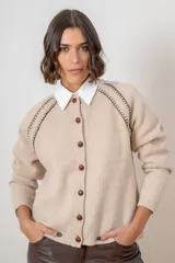 Saco de punto color beige, con cierre frontal de botones marrones y detalle de costuras decorativas oscuras en los hombros. Presenta un diseño clásico con cuello redondo.
