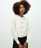 Cardigan de punto color blanco, con cuello redondo, manga larga y cierre frontal con botones metálicos decorativos en forma de flor. Presenta un diseño de corte regular con acabados acanalados en puños y ruedo.