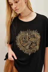 Remera negra de corte holgado con cuello redondo y mangas cortas. Presenta un aplique central en forma de corazón bordado con hilos y cuentas doradas, con un diseño detallado de aves en su interior.