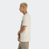 Remera de manga corta color crema con un estampado gráfico de corredores en colores vibrantes (amarillo, azul, rosa, celeste). Incluye el logo de New Balance en la parte inferior del estampado.