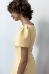 Vestido corto color amarillo claro, con cuello barco, mangas cortas abullonadas y pliegues en la parte delantera. Presenta falsos bolsillos de vivo y costuras marcadas. Cierre en la espalda con cremallera oculta.