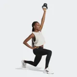 Musculosa deportiva Adidas de corte holgado (boxy) y largo recortado (cropped), color beige claro, con cuello redondo y aberturas amplias en las sisas. Presenta el logo de Adidas Badge of Sport en blanco en el pecho.