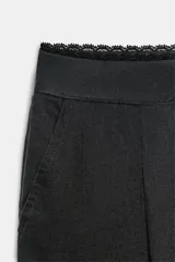 Pantalón de lino negro de tiro medio, con pernera recta, pinzas delanteras y detalle de puntilla en la cintura. Cuenta con bolsillos frontales y cierre con cremallera y botón.