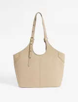 Cartera tipo shopper color beige, con diseño de costura central vertical y detalles de tachas metálicas doradas en los laterales superiores. Cuenta con correas ajustables mediante hebillas metálicas y logo grabado en la parte inferior.